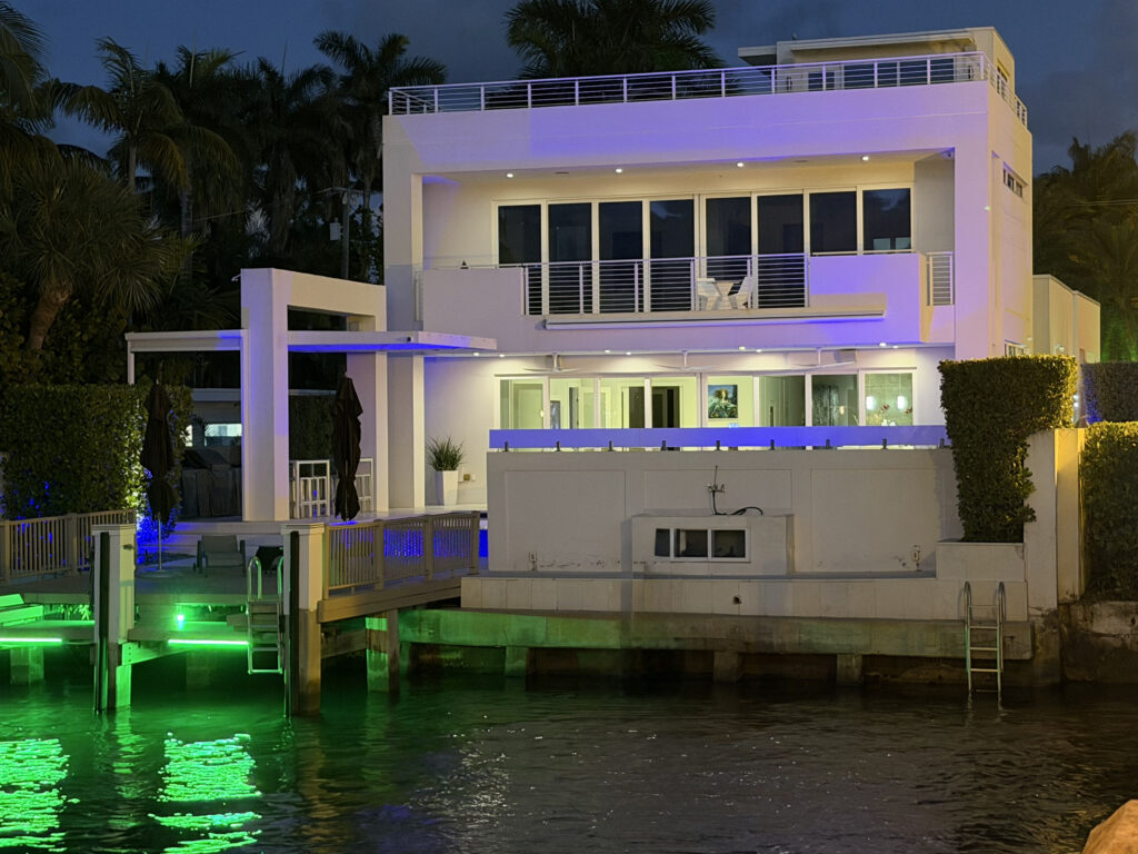 A modern waterfront house illuminated at night with a dock and palm trees.